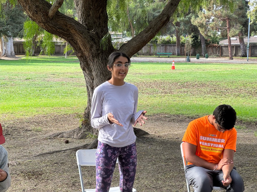 Outdoor community gathering under a tree: instructor speaking with a participant at a Brave Buds event